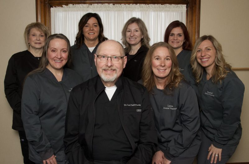 A group portrait of a dental team consisting of eight professionals in dark scrubs and lab coats smiling in an office setting.